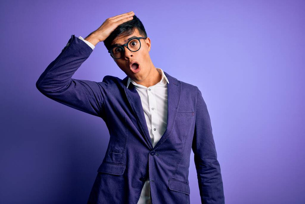 Young handsome business man wearing jacket and glasses over isolated purple background surprised with hand on head for mistake, remember error. Forgot, bad memory concept. - Фото, изображение