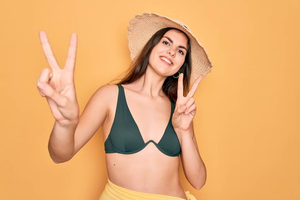Young beautiful girl wearing swimwear bikini and summer sun hat over yellow background smiling looking to the camera showing fingers doing victory sign. Number two. - Foto, Imagem
