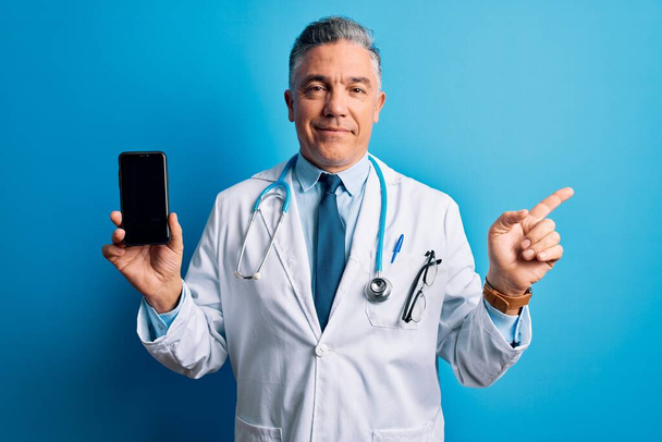 Middle age handsome grey-haired doctor man holding smartphone showing screen very happy pointing with hand and finger to the side - Zdjęcie, obraz