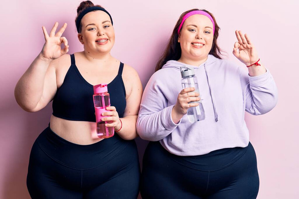 Young plus size twins wearing sportswear drinking bottle of water doing ok sign with fingers, smiling friendly gesturing excellent symbol  - Foto, immagini