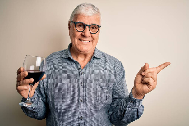 Ältere gut aussehende Mann hoary trinken Glas Rotwein über isolierten weißen Hintergrund sehr glücklich zeigt mit Hand und Finger zur Seite - Foto, Bild