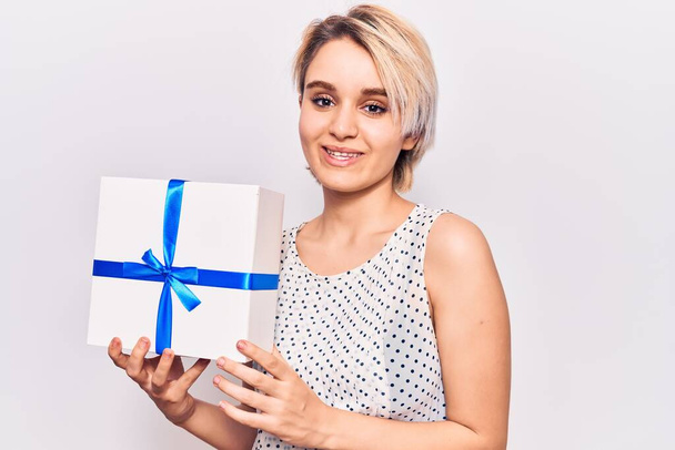 Young beautiful blonde woman holding gift looking positive and happy standing and smiling with a confident smile showing teeth  - Photo, Image