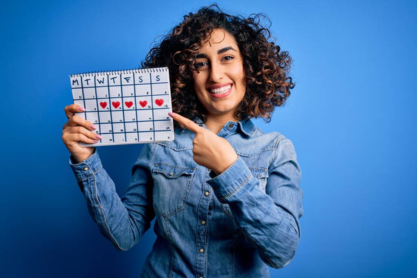 Junge schöne lockige arabische Frau mit Zeitrahmen-Kalender zur Kontrolle des Menstruationszyklus sehr glücklich zeigt mit Hand und Finger - Foto, Bild