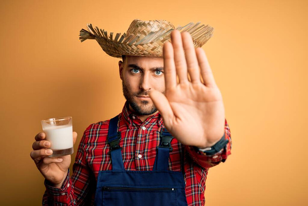 Jonge boerenman op het platteland draagt hoed drinken biologische melk van boerderij met open hand doen stopbord met serieuze en zelfverzekerde uitdrukking, verdediging gebaar - Foto, afbeelding