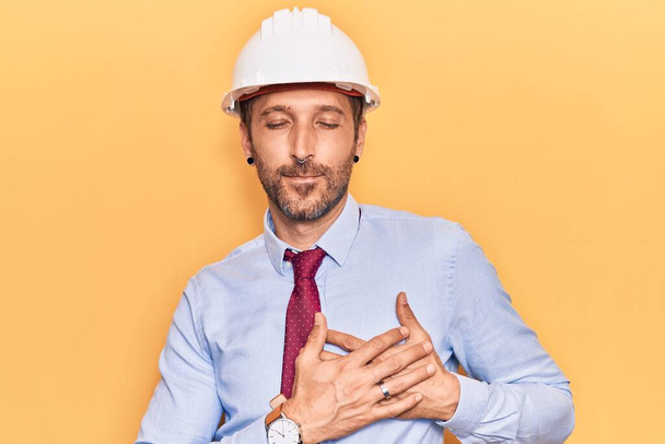 Mladý pohledný muž v architektově klobouku, s rukama na prsou, oči zavřené vděčným gestem na tváři. zdravotní koncepce.  - Fotografie, Obrázek