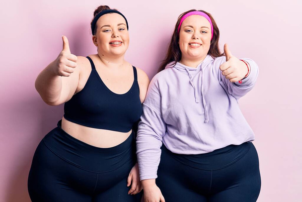 Young plus size twins wearing sportswear approving doing positive gesture with hand, thumbs up smiling and happy for success. winner gesture.  - Fotografie, Obrázek