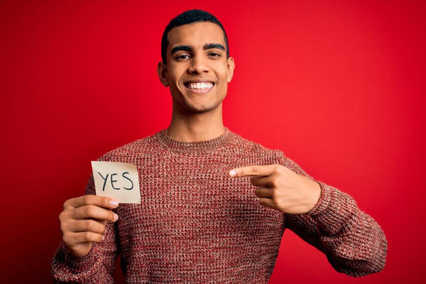 Young handsome african american man holding reminder paper with yes message very happy pointing with hand and finger - Zdjęcie, obraz