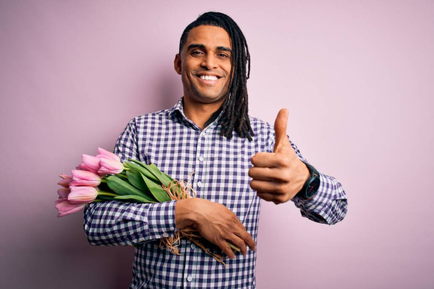 Giovane afroamericano afro romantico uomo con dreadlocks tenendo bouquet di tulipani rosa facendo felice pollice in alto gesto con la mano. Approvazione espressione guardando la fotocamera che mostra il successo. - Foto, immagini