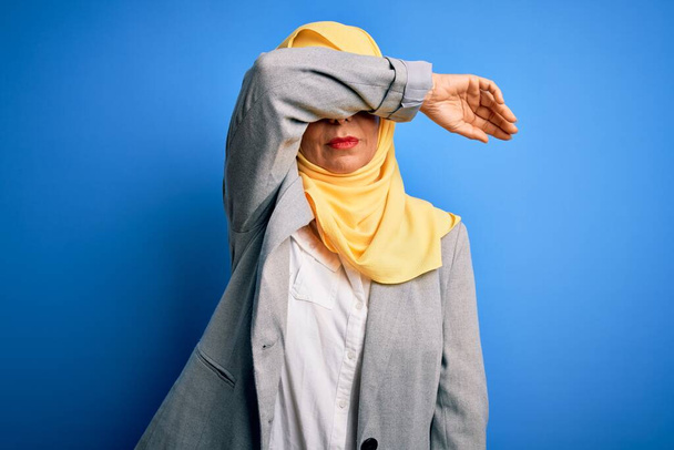 Orta yaşlı esmer iş kadını, geleneksel Müslüman tesettüre bürünmüş mavi arka planda, gözleri kolla kaplanmış, ciddi ve üzgün görünüyor. Görme, saklama ve reddetme konsepti - Fotoğraf, Görsel