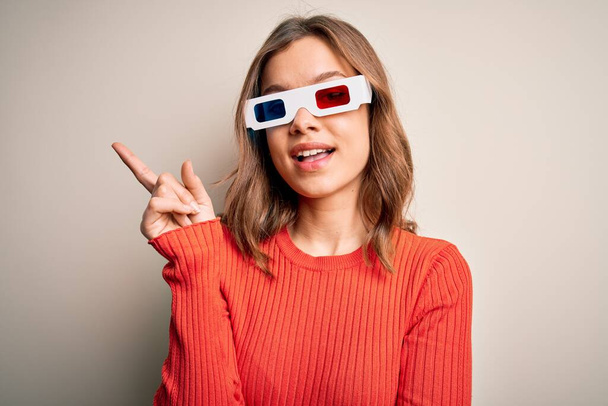 Young blonde girl wearing 3d cinema glasses over isolated background with a big smile on face, pointing with hand and finger to the side looking at the camera. - Zdjęcie, obraz