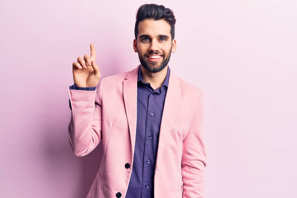 Young handsome man with beard wearing elegant jacket showing and pointing up with finger number one while smiling confident and happy.  - Photo, Image