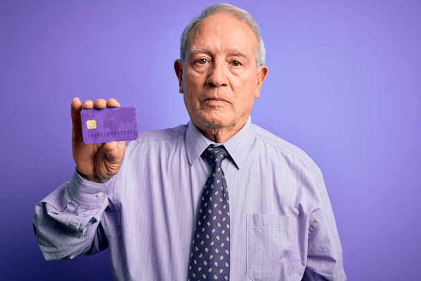 Senior grey haired business man holding credit card over purple background with a confident expression on smart face thinking serious - Фото, зображення