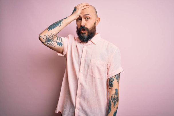 Handsome bald man with beard and tattoo wearing casual shirt over isolated pink background surprised with hand on head for mistake, remember error. Forgot, bad memory concept. - 写真・画像