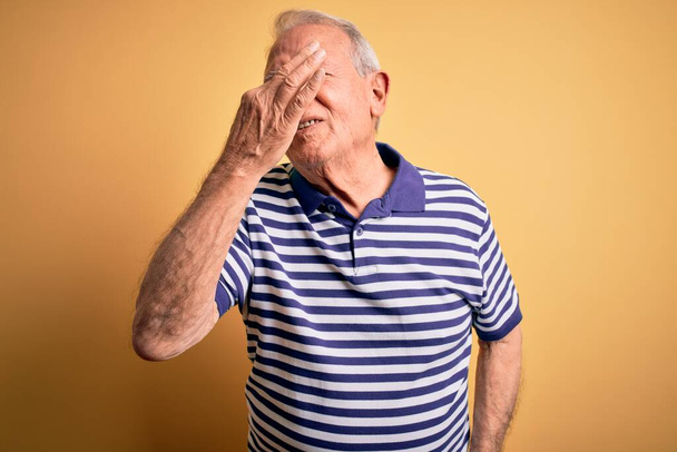 Grey haired senior man wearing casual navy striped t-shirt standing over yellow background smelling something stinky and disgusting, intolerable smell, holding breath with fingers on nose. Bad smell - Фото, изображение