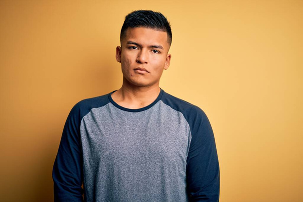 Young handsome latin man wearing casual t-shirt standing over yellow background Relaxed with serious expression on face. Simple and natural looking at the camera. - Foto, Bild