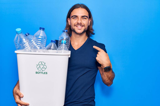 Jeune homme beau tenant corbeille de recyclage avec des bouteilles en plastique pointant du doigt un sourire heureux et fier  - Photo, image