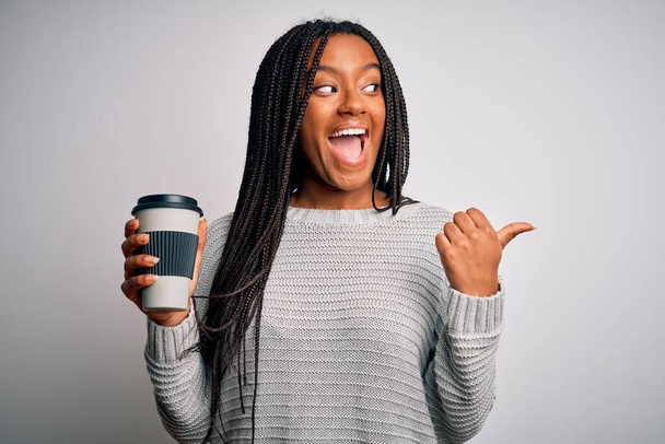 Young african american girl drinking coffe on a take away plastic cup over isolated background pointing and showing with thumb up to the side with happy face smiling - Photo, Image
