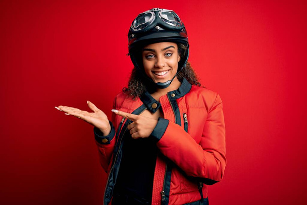 Young african american motorcyclist girl wearing moto helmet and glasses over red background amazed and smiling to the camera while presenting with hand and pointing with finger. - Foto, Bild