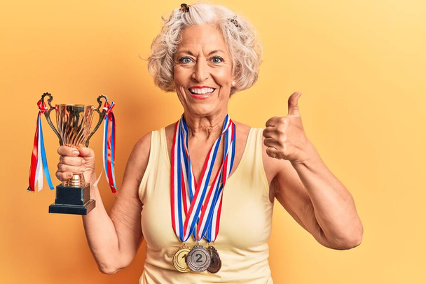 Senior grey-haired woman holding champion trophy wearing medals smiling happy and positive, thumb up doing excellent and approval sign  - Valokuva, kuva