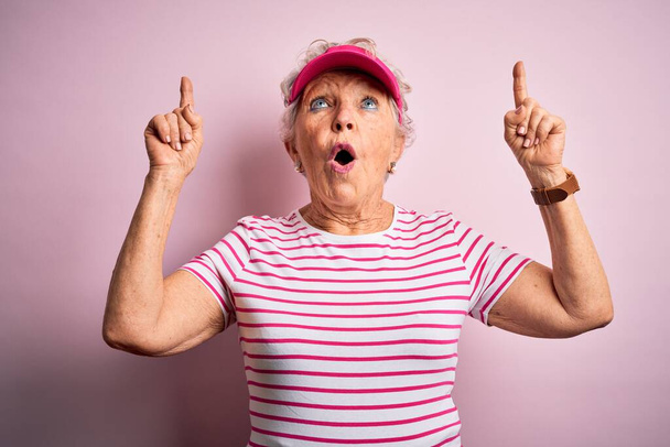 Senior belle femme sportive portant une casquette de sport debout sur fond rose isolé étonné et surpris de regarder vers le haut et pointant avec les doigts et les bras levés. - Photo, image