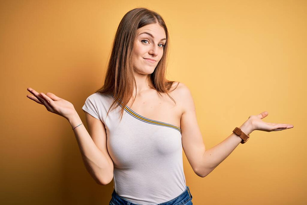 Νεαρή όμορφη κοκκινομάλλα γυναίκα φορώντας casual t-shirt πάνω από απομονωμένο κίτρινο φόντο ανίδεο και σύγχυση έκφραση με τα χέρια και τα χέρια ψηλά. Έννοια αμφιβολίας. - Φωτογραφία, εικόνα