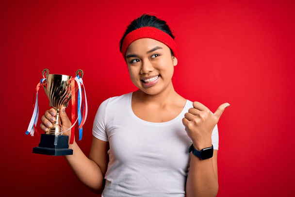 Joven campeón asiático chica ganar trofeo taza de pie sobre aislado rojo fondo apuntando y mostrando con el pulgar hasta el lado con feliz cara sonriendo
 - Foto, Imagen