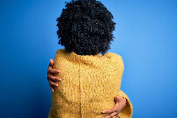 Junge schöne afroamerikanische Afrofrau mit lockigem Haar, die einen gelben lässigen Pullover trägt und sich glücklich und positiv von hinten umarmt. Selbstliebe und Selbstfürsorge - Foto, Bild