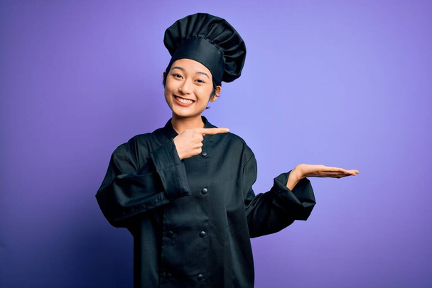 若いです美しい中国のシェフの女性は、手で提示しながら、カメラに驚いて、笑顔と指で指す紫色の背景に炊飯器の制服と帽子を身に着けて. - 写真・画像