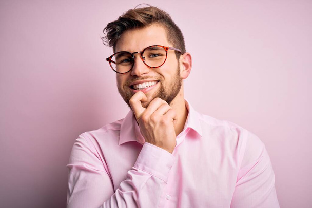 Jonge knappe blonde man met baard en blauwe ogen met roze hemd en bril die zelfverzekerd naar de camera kijkt met een glimlach met gekruiste armen en hand op de kin. Positief denken. - Foto, afbeelding