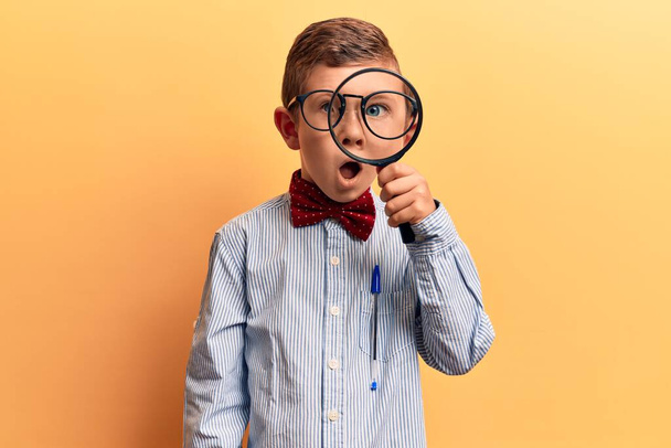 Cute blond kid wearing nerd bow tie and glasses holding magnifying glass scared and amazed with open mouth for surprise, disbelief face  - Fotoğraf, Görsel