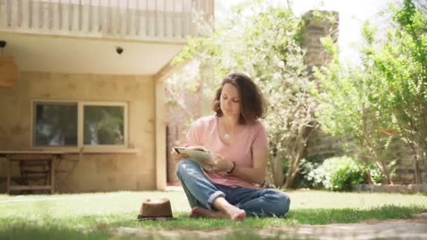 Young happy woman sits in hat on a lawn in country house, writing diary - Séquence, vidéo