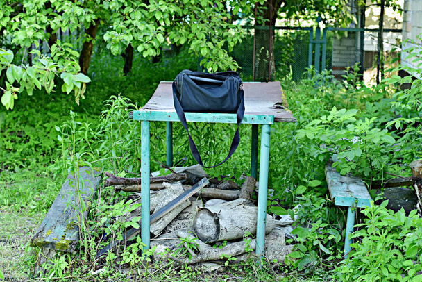 В заросшем травой саду стоит металлический стол, на котором стоит мешок, в заросшем саду - сумка. Высокое качество фото
 - Фото, изображение