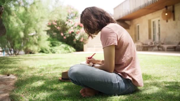 Młoda szczęśliwa kobieta siedzi na trawniku w wiejskim domu i pisze pamiętnik - Materiał filmowy, wideo