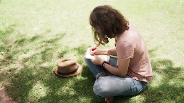  Jeune femme heureuse assise dans un chapeau sur une pelouse et prend des notes dans un carnet. - Séquence, vidéo