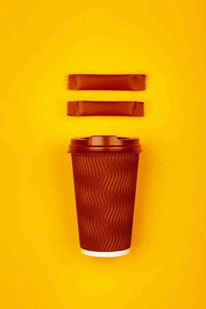 Κοντινό πλάνο ένα τσαλακωμένο καφέ χάρτινο κύπελλο takeaway μιας χρήσης και δύο φακελάκι ζάχαρη stick πάνω από ζωντανό κίτρινο φόντο, επίπεδη lay, υπερυψωμένη πάνω όψη, ακριβώς πάνω από - Φωτογραφία, εικόνα