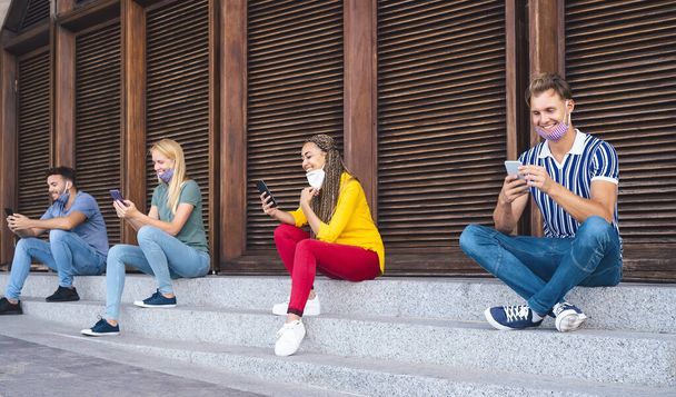 Молодые люди используют мобильные телефоны, сохраняя социальную дистанцию на открытом воздухе - Мультирасовые друзья весело смотрят на смартфоны приложения социальных сетей - Технологии и молодежное поколение во время вспышки
 - Фото, изображение