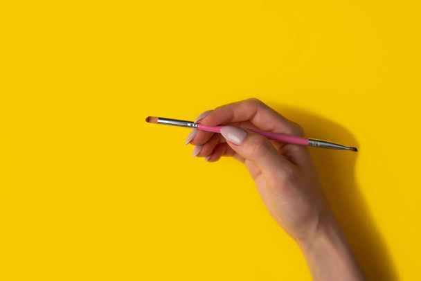 Female hand with a brush on a yellow background, top view.Manicure concept, flat lay - Foto, afbeelding