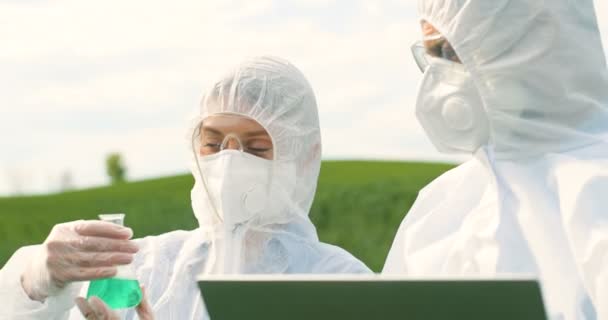 Close up of Caucasian female and male farmers researchers holding test tube with chemicals pesticides and laptop computer in green field. Biologists co-workers working and talking in wheat margin. - Footage, Video