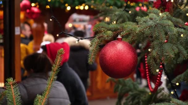 Großaufnahme einer roten Weihnachtskugel, die an einem natürlichen Neujahrsbaum auf einem Stadtfest hängt, Menschenmenge auf einem festlichen Platz. - Filmmaterial, Video