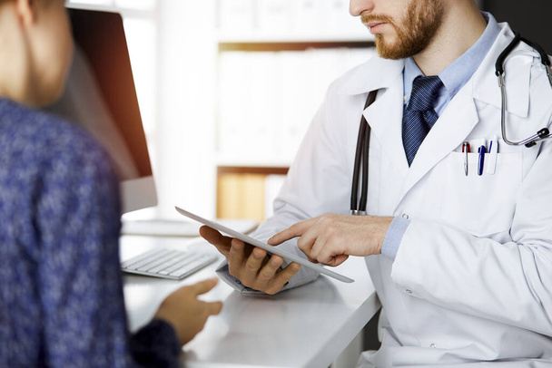 Unknown red-bearded doctor and patient-woman discussing current health examination while sitting and using tablet computer in sunny clinic, close-up. Medicine concept - Foto, Bild