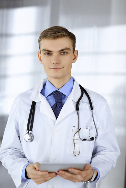 Young doctor man is smiling cheerfully at camera, while standing in a hospital office, holding a computer tablet in his hands. Perfect medical service in clinic. Happy future in medicine and - Фото, зображення