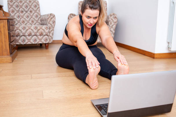 Plus Size Frau, die ihren Körper bei einem Fitnesskurs zu Hause dehnt. Sie benutzt ihren Computer, um sich Videostunden anzusehen. - Foto, Bild