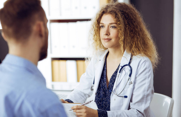 Médico femenino y paciente masculino discutiendo el examen de salud actual mientras está sentado en una clínica soleada. Concepto de medicina - Foto, Imagen