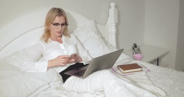 Businesswoman in eye glasses read text message on smartphone working on laptop. Business woman relaxes in bed after a long flight. Business concept. Prores 422 - Séquence, vidéo
