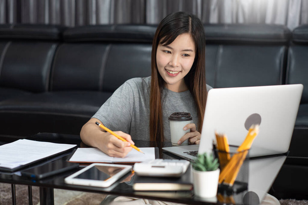 Vonzó, fiatal, gyönyörű ázsiai nő, aki laptoppal és dokumentumokkal dolgozik, kávét iszik a nappaliban, szabadúszóként, e-coachingként dolgozik, távolról vagy otthonról dolgozik. - Fotó, kép