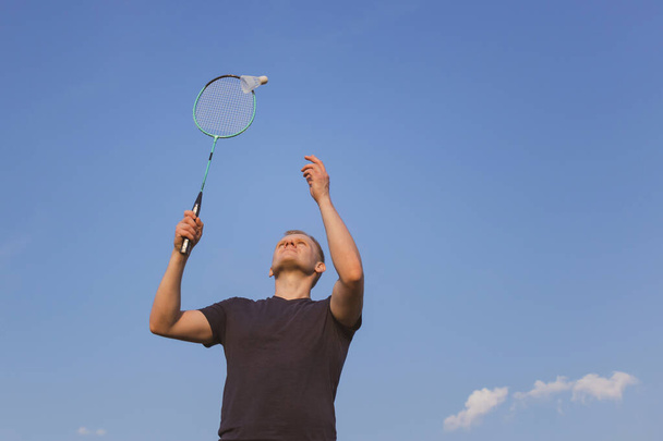 Young Caucasian man plays badminton on a background of blue sky. The concept of an amateur game of badminton, outdoor activities. Copyspace, bottom view. - Photo, Image