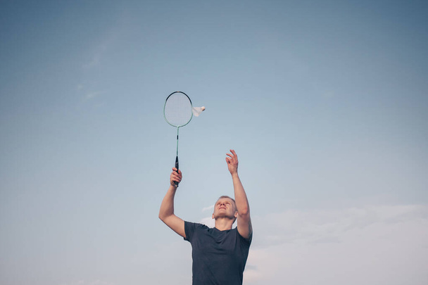 Młody biały mężczyzna gra badmintona na tle błękitnego nieba. Koncepcja amatorskiej gry badmintona, zajęcia na świeżym powietrzu. Copyspace, widok z dołu - Zdjęcie, obraz