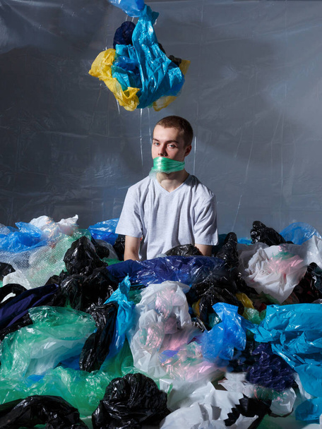 Widok pionowy. Młody mężczyzna siedzący, przywiązany do ust plastikową torbą, otoczony plastikowymi torbami, patrzący w kamerę. - Zdjęcie, obraz
