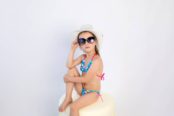 a little girl of 5-6 years old is sitting in a swimsuit with glasses in a hat on a white isolated background, a place for text - Фото, изображение