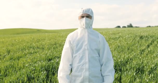 Portret van de blanke mannelijke boer ecoloog in wit beschermend kostuum, masker en bril staan in het groene veld in de zomer. Mens wetenschapper en bioloog in de marge met eco oogst. - Video